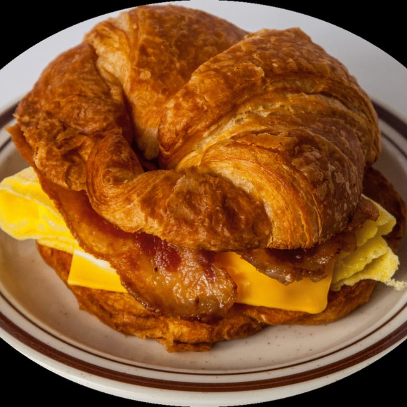 A breakfast croissant sandwich with bacon, egg, and cheese on a white plate.