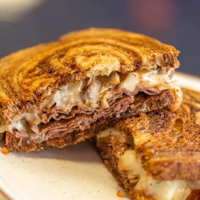 Close-up of a grilled sandwich, cut in half, revealing melted cheese and thinly sliced meat.