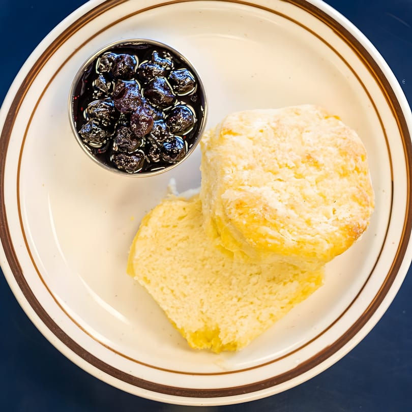 Fluffy biscuits and a small metal cup of dark blueberry jam on a white plate with brown stripes.